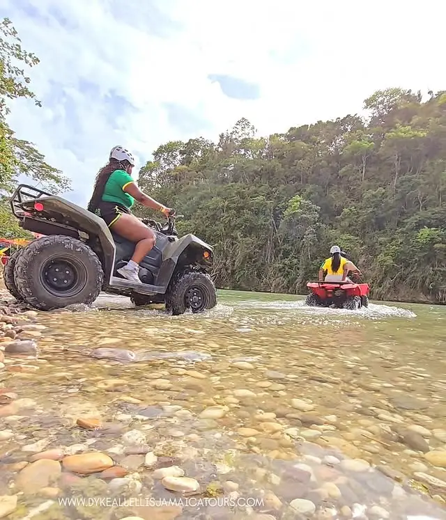 Two guests riding ATVs across a shallow rocky river in the Jamaican bush